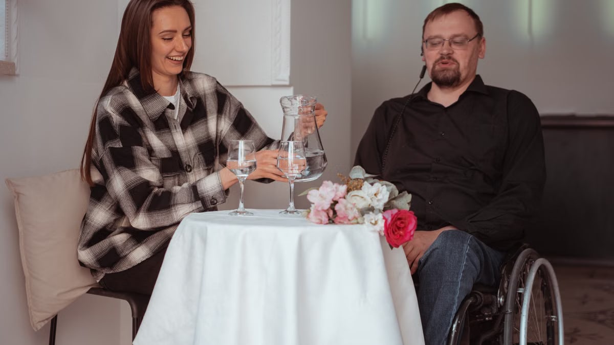 A couple seated together, one in a wheelchair, sharing a moment over drinks indoors.